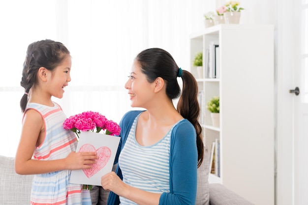 Menina asiática sorridente preparar um buquê de cravo rosa e cartão de amor dando presente para linda mãe no dia das mães em casa no sofá da sala olhando uns aos outros com copyspace.