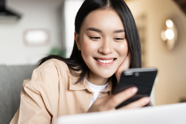 Menina asiática sorridente usando um celular, trabalhando em um laptop e verificando a mensagem no celular