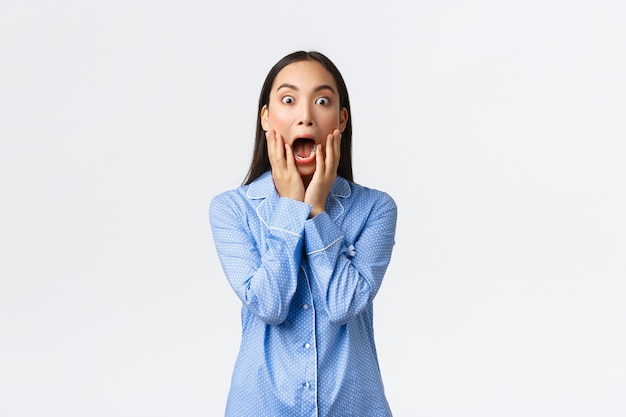 Menina asiática surpresa e chocada de pijama azul, gritando de espanto e espanto, segurando a boca aberta e olhando para a câmera impressionada, fundo branco de pé.