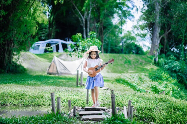 Menina asiática tocando ukulele ou guitarra havaiana no parque enquanto acampa no verão