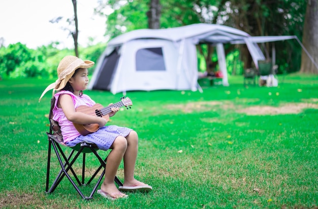 Menina asiática tocando ukulele ou guitarra havaiana no parque enquanto acampa no verão