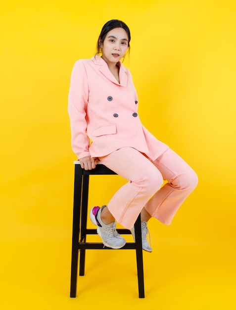 Menina asiática travessa com calça e jaqueta rosa pastel, senta-se vagarosamente na cadeira alta, posando de modelo para a moda adulta. Retrato relaxante para roupas modernas e terno para roupa feminina.
