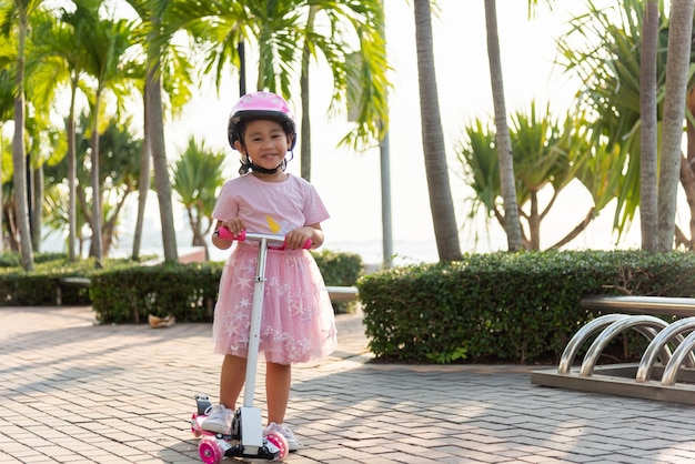 Menina asiática usa capacete seguro jogando prancha rosa na estrada no parque ao ar livre no verão