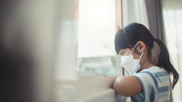 Menina asiática usando máscara para proteger pm2.5 e Covid-19. Criança doente Meninas olham para a janela com saudade na rua. Copie o espaço. criança tristeza em casa em isolamento