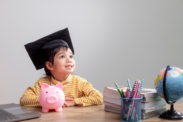 Menina asiática usando um chapéu de formatura com um cofrinho rosa Economizando dinheiro investindo no futuro