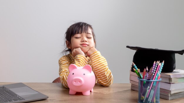 Menina asiática usando um chapéu de formatura com um cofrinho rosa Economizando dinheiro investindo no futuro