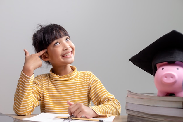 Menina asiática usando um chapéu de formatura com um cofrinho rosa Economizando dinheiro investindo no futuro