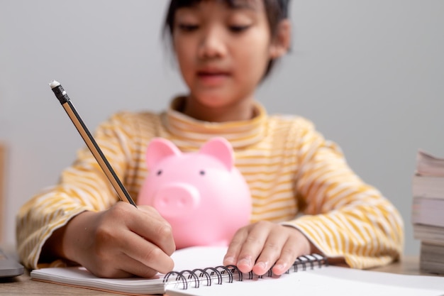 Menina asiática usando um chapéu de formatura com um cofrinho rosa Economizando dinheiro investindo no futuro