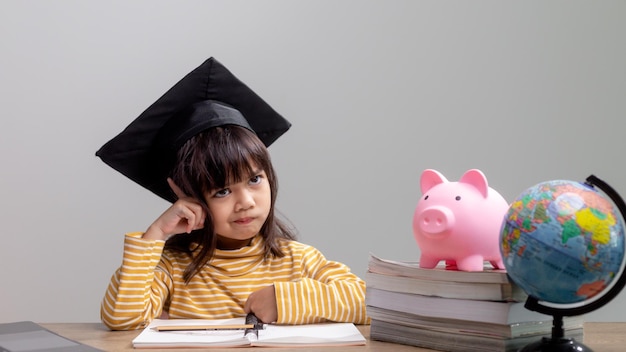 Menina asiática usando um chapéu de formatura com um cofrinho rosa Economizando dinheiro investindo no futuro