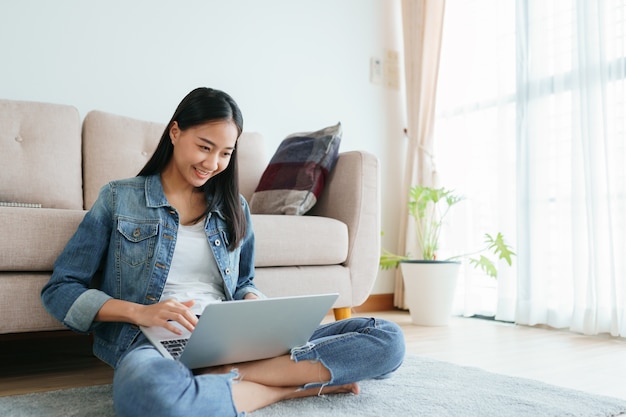 Foto menina asiática vestindo jeans usando um laptop enquanto está sentado no chão em casa.