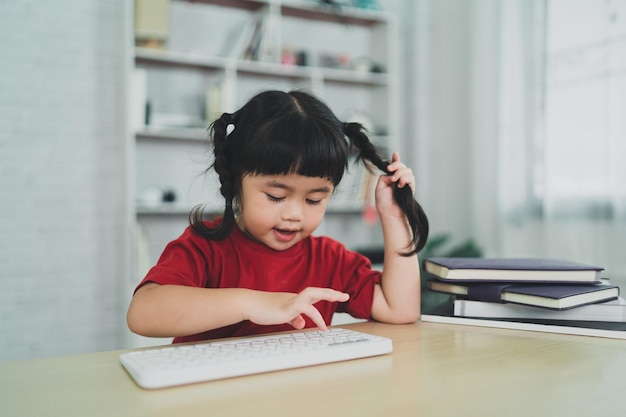 Menina asiática vestindo uma camiseta vermelha usa teclado sem fio e estuda on-line na mesa de madeira na sala de estar em casa Educação aprendendo on-line a partir do conceito de casa