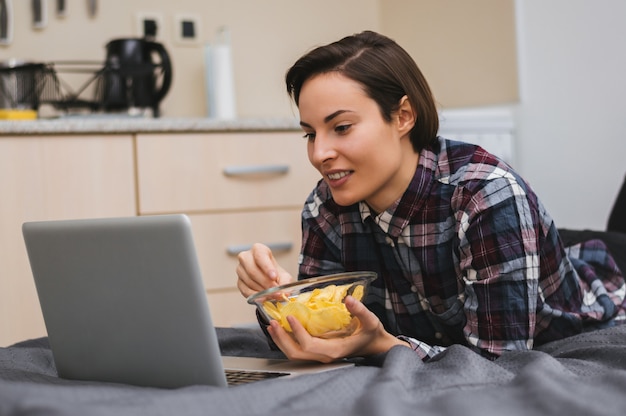 Menina assistindo a um filme no computador e deitado na cama, comendo batatas fritas