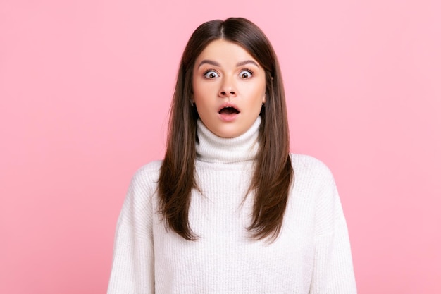 Menina assustada chocada olhando para a câmera com a boca aberta e olhos grandes, vê algo surpreendente, vestindo suéter branco estilo casual. Tiro de estúdio interior isolado no fundo rosa.