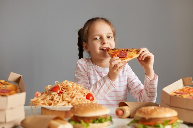 Menina atraente com fome e otimista com tranças sentadas à mesa com junk food isolada sobre fundo cinza mordendo uma grande fatia de pizza desfrutando de um jantar de bom apetite