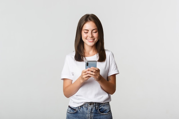 Foto menina atraente sorridente usando telefone celular e olhando para a tela satisfeito.