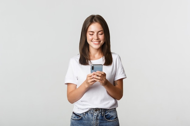 Menina atraente sorridente usando telefone celular e olhando para a tela satisfeito.