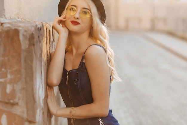 Menina atrativa loura nos óculos de sol e no chapéu à moda no sol do verão urbano