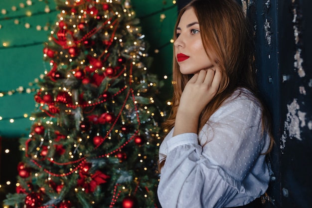 Menina atrativa nova na roupa festiva no fundo de uma árvore de Natal