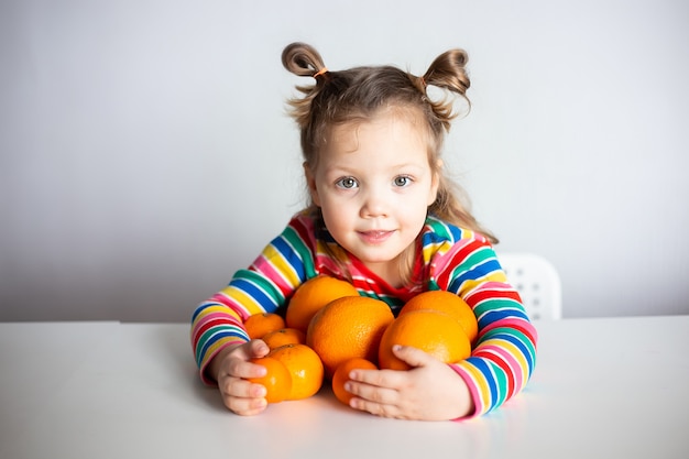 Menina, bebê de 3 anos, com um penteado de rabo de cavalo em uma jaqueta listrada colorida e colorida abraça um monte de appelsins