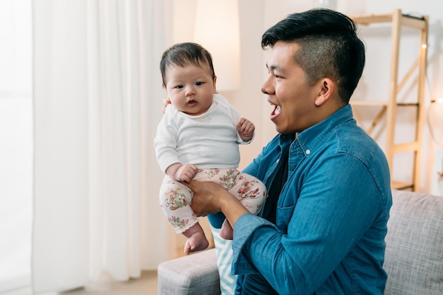 Menina bebê recém-nascido bonito asiático está sentado nas mãos de seu pai com inocência enquanto seu pai está olhando e brincando com ela em uma sala de estar brilhante em casa.