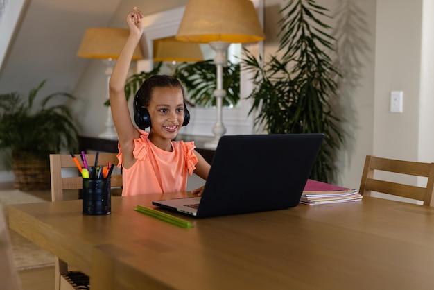 Menina biracial feliz usando fones de ouvido e laptop para a lição on-line em casa levantando a mão