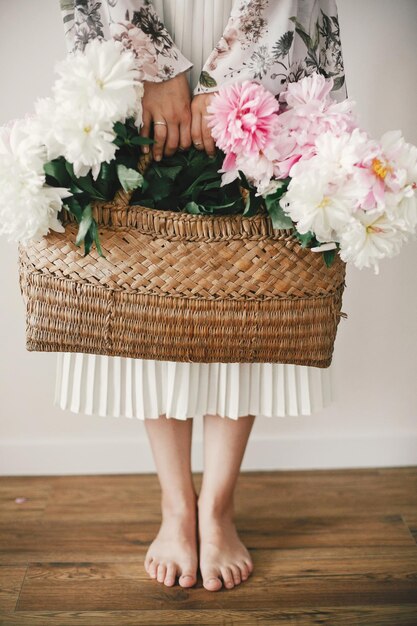 Menina Boho segurando peônias rosa e brancas na cesta rústica e de pé descalço no chão de madeira Mulher elegante hipster em vestido floral boêmio com flores de peônia Dia Internacional da Mulher