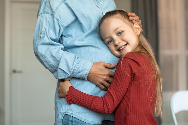 Foto menina bonita abraçando a barriga grande da mãe grávida e sorrindo para o garoto da câmera se relacionando com a mãe grávida
