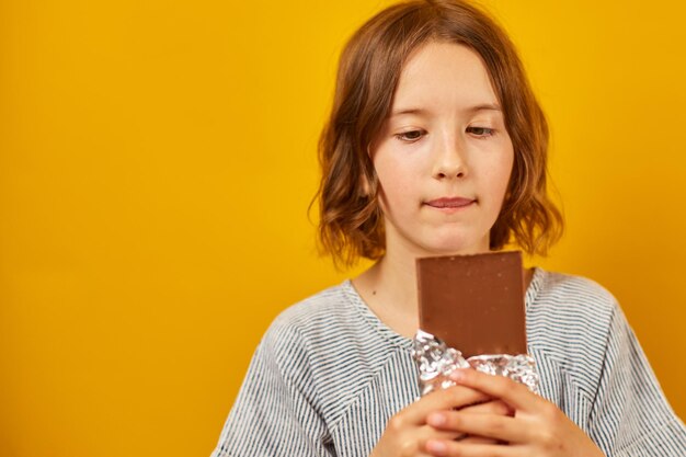 Foto menina bonita adolescente com uma barra de chocolate isolada no fundo amarelo do estúdio, aproveitando o espaço de cópia de sobremesa