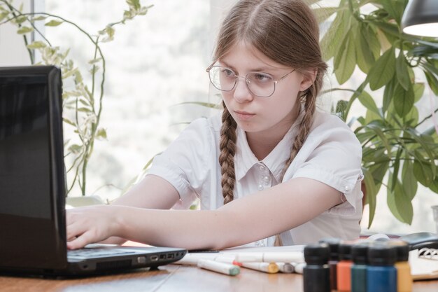 Menina bonita adolescente digitando em um laptop e fazendo lição de casa sentado atrás da mesa em casa. Linda menina de óculos, aprendendo on-line com um laptop, ensino à distância, autoeducação