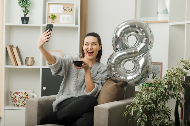 Menina bonita animada piscando no dia da mulher feliz segurando uma taça de vinho, tirar uma selfie sentada na poltrona da sala de estar