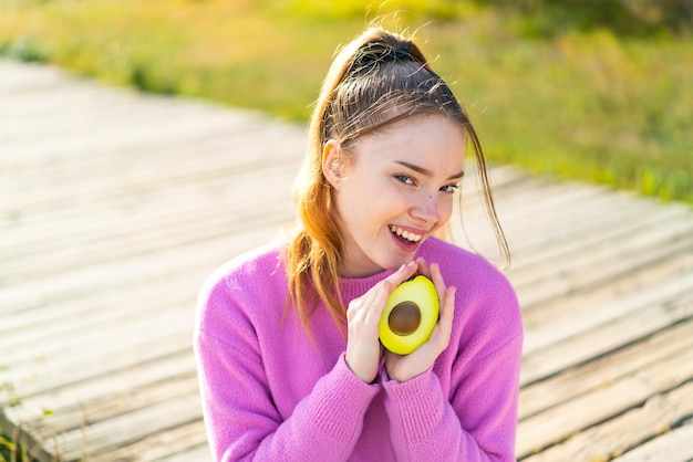 Menina bonita ao ar livre segurando um abacate com expressão de surpresa