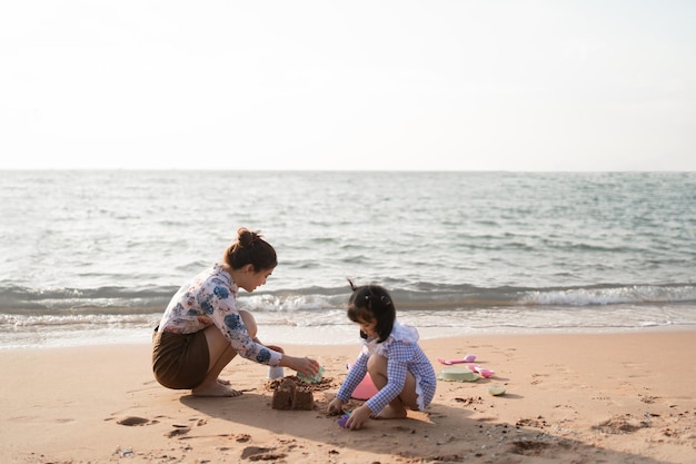 Menina bonita asiática e sua mãe brincando ou fazendo castelo de areia ou cavando com areia na praia tropical Crianças com lindo céu azul de areia do mar Crianças felizes em férias à beira-mar na praia