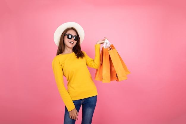 Menina bonita asiática que guarda os sacos alaranjados da compra que olham afastado no fundo cor-de-rosa, conceito colorido da compra.