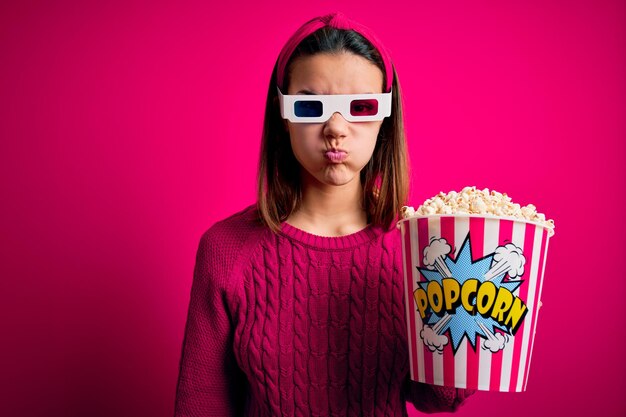 Menina bonita assistindo filme usando óculos 3d comendo caixa com pipocas estufando as bochechas com cara engraçada Boca inflada com expressão louca de ar