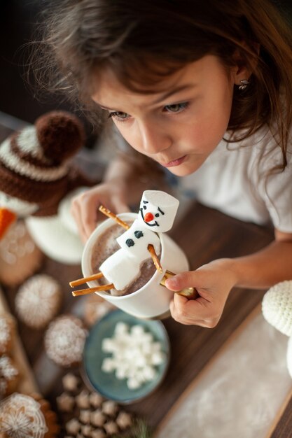 Menina bonita brinca com bonecos de neve de malha e come pão de mel e bebe chocolate com marshmallow. Cozinha e sala de jantar elegantes em casa. Conceito de preparação de natal