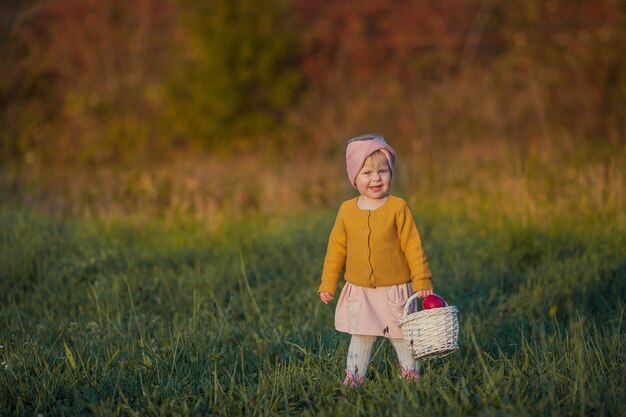 Menina bonita caminha no jardim outono, segura uma cesta com maçãs vermelhas. Retrato de uma menina feliz com roupas brilhantes de outono. Outono quente e brilhante.