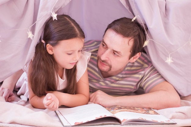 Menina bonita caucasiana e o pai dela deitado com um livro na cabana e lendo