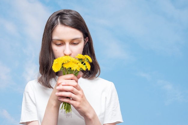 Menina bonita caucasiana inala o cheiro de dente de leão amarelo contra um céu azul com nuvens, copie o espaço. A menina recebeu um buquê de flores silvestres de presente. Flores de verão como um presente.