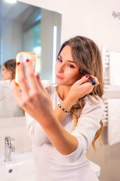 Menina bonita caucasiana se maquiando no banheiro, olhando no espelho, foto vertical