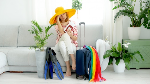 Menina bonita chateada com um chapéu amarelo fica em casa e planeja uma viagem de férias. Mala e nadadeiras para mergulho.