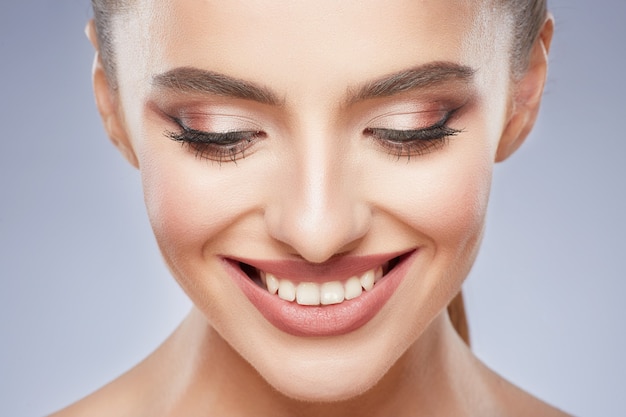 Menina bonita com cabelo castanho preso atrás, pele limpa, fresca, olhos grandes e ombros nus, posando no fundo cinza do estúdio, olhando para baixo e sorrindo, retrato de modelo.