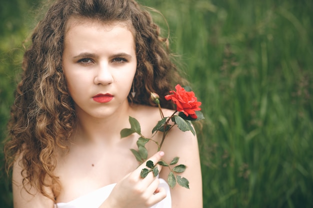 Menina bonita com cabelo comprido e rosa vermelha nas mãos em pé em um campo verde