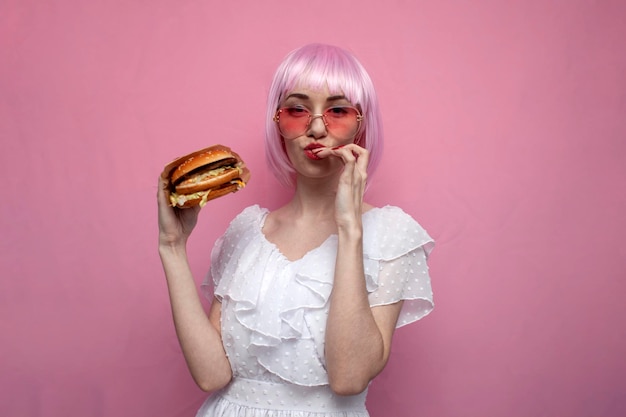 Menina bonita com cabelo curto rosa segura um grande hambúrguer nas mãos e lambe os lábios, uma mulher mostra um hambúrguer