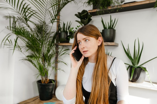 Menina bonita com cabelo loiro falando ao telefone dentro de casa com plantas e parede branca olhando para longe