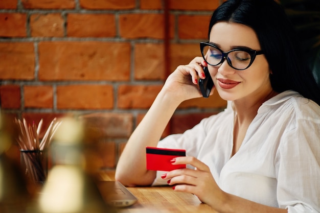 Menina bonita com cabelo preto, usando óculos, sentado no café com laptop, telefone celular, cartão de crédito e xícara de café, conceito freelance, compras online, camisa branca.