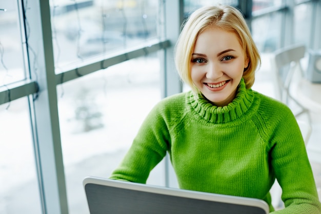 Menina bonita com cabelos claros, suéter verde, sentado no café com laptop, retrato, conceito freelance, compras online.