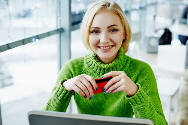 Menina bonita com cabelos claros, suéter verde, sentado no café com laptop, retrato, conceito freelance, compras online.