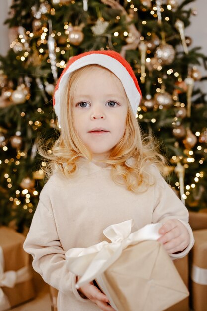 Menina bonita com chapéu de papai noel sob a árvore de natal com caixa de presente. boas festas, ano novo. aconchegante noite de inverno quente em casa. hora do natal