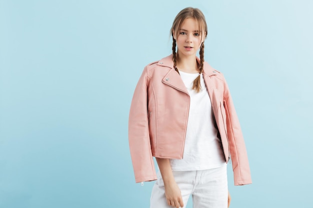 Menina bonita com duas tranças na jaqueta de couro rosa e camiseta branca sonhadoramente olhando na câmera sobre fundo azul isolado