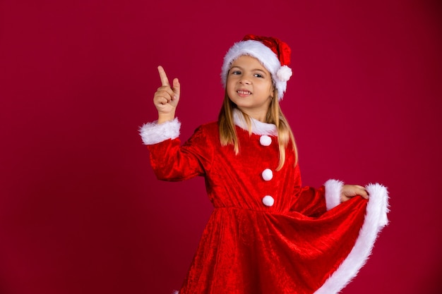 Foto menina bonita com fantasia de papai noel e chapéu de inverno. retrato de criança segurando o vestido vermelho com a mão e apontando o dedo. isolado na parede vermelha com espaço livre.